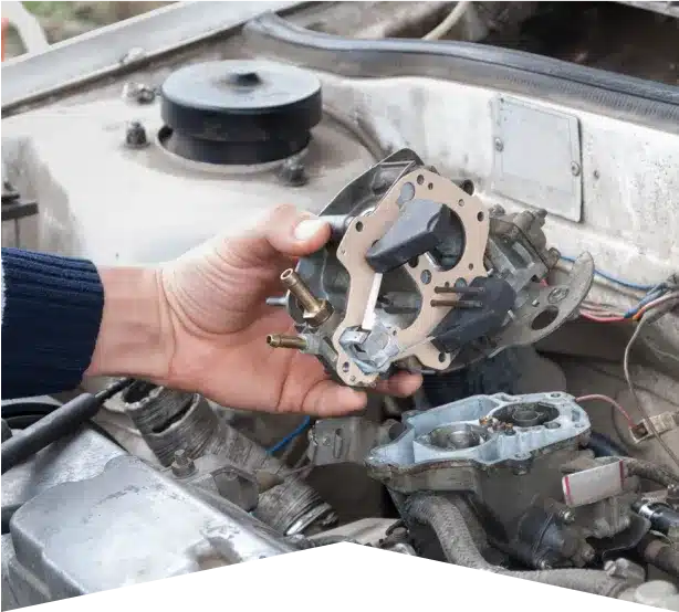Carburetor Services in Clive, IA At Westside Auto Pros. A person’s hand holds a carburetor above the open engine bay of a car, preparing to install or remove the part. Various engine components and wiring are visible in the background.