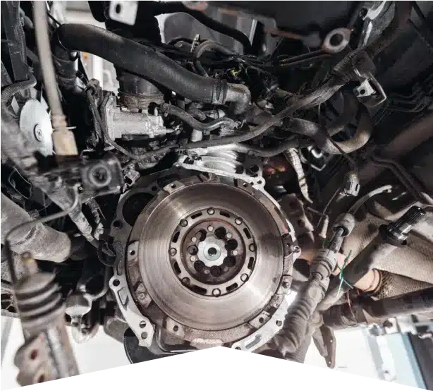 Clutch Repair in Clive, IA At Westside Auto Pros. A close-up view of a car engine’s exposed flywheel and surrounding mechanical components, with various wires, hoses, and metal parts visible in a garage setting.