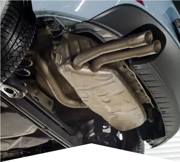 Exhaust System Repair in Clive, IA At Westside Auto Pros. Close-up view of a cars undercarriage showing the exhaust system with dual tailpipes and a muffler, next to a rear tire. The car appears to be lifted for inspection or repair.