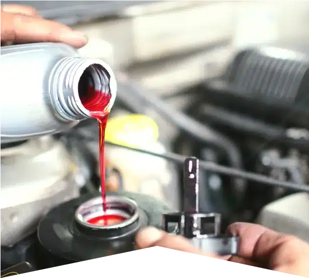 Fluid Services in Clive, IA At Westside Auto Pros. A person pours red fluid from a bottle into a car engine compartment, likely adding transmission or power steering fluid, with car parts visible in the background.
