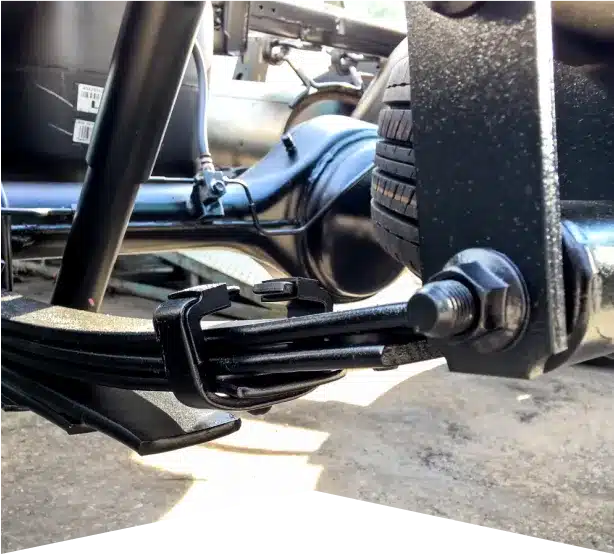 Suspension & Steering Repair in Clive, IA At Westside Auto Pros. Close-up view of a vehicle’s rear suspension system, showing leaf springs, a solid axle, U-bolts, and part of a tire, with a blurred background of the vehicle’s undercarriage.