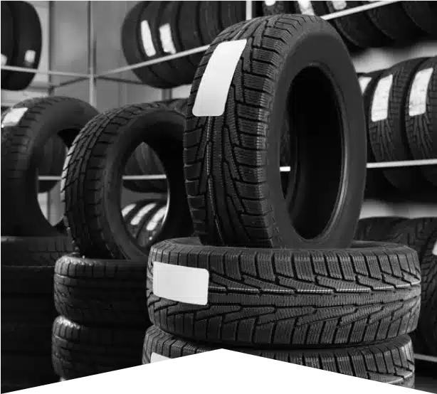Tire Sales & Services in Clive, IA At Westside Auto Pros. Stacks of new car tires with labels are arranged on shelves and the floor in a tire shop. The image is in black and white, highlighting the tread patterns and clean appearance of the tires.