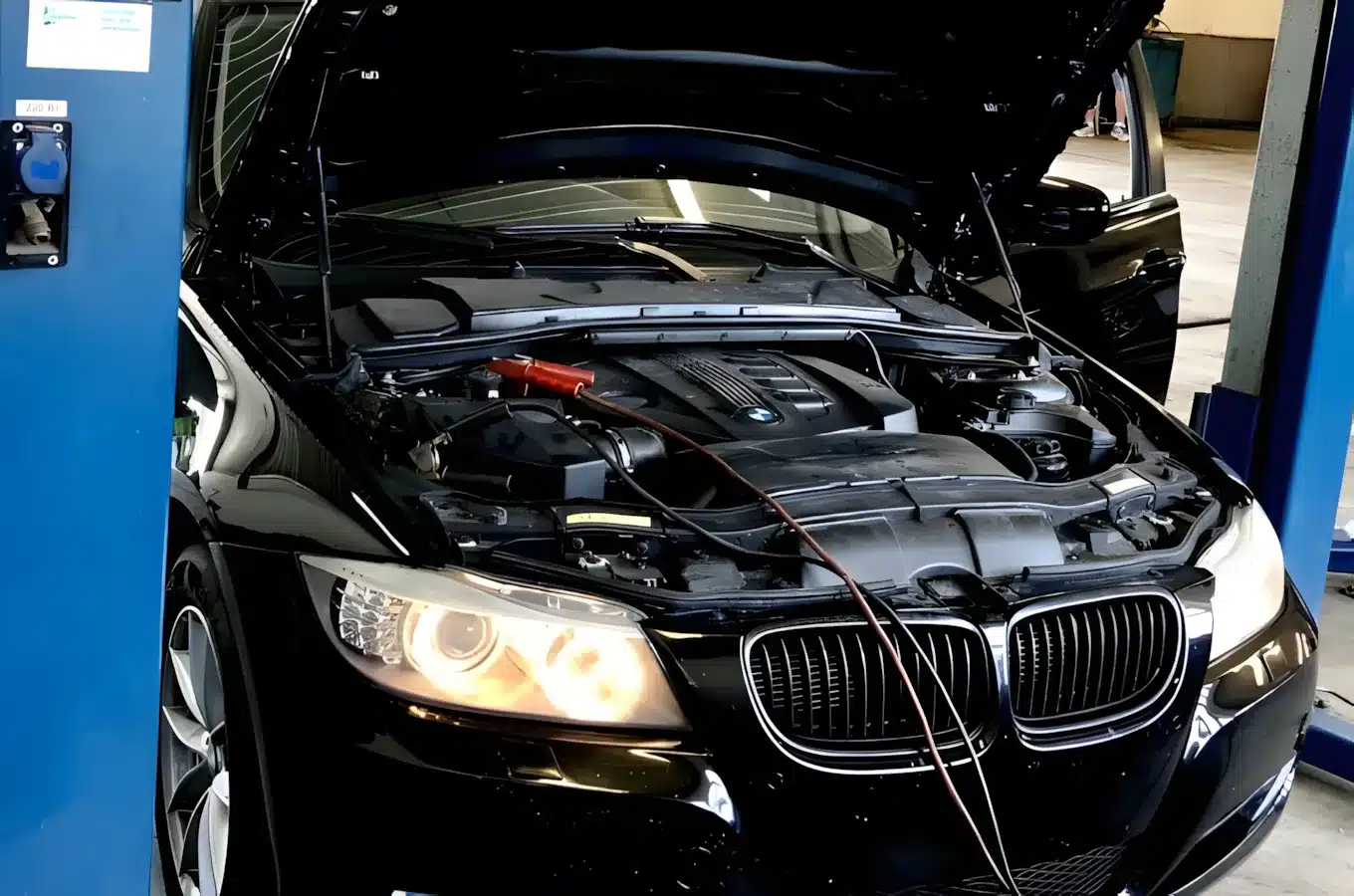 A black car with its hood open is being serviced. Jumper cables are attached to the engine, and the left headlight is on. The car is parked in a garage or workshop.
