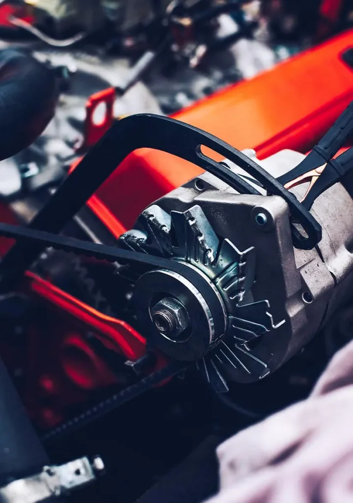 Alternator Repair & Service in Clive, IA At Westside Auto Pros. Close-up of a car engine showing a metal alternator with a black drive belt looped around a pulley, set against a bright orange engine component in the background.