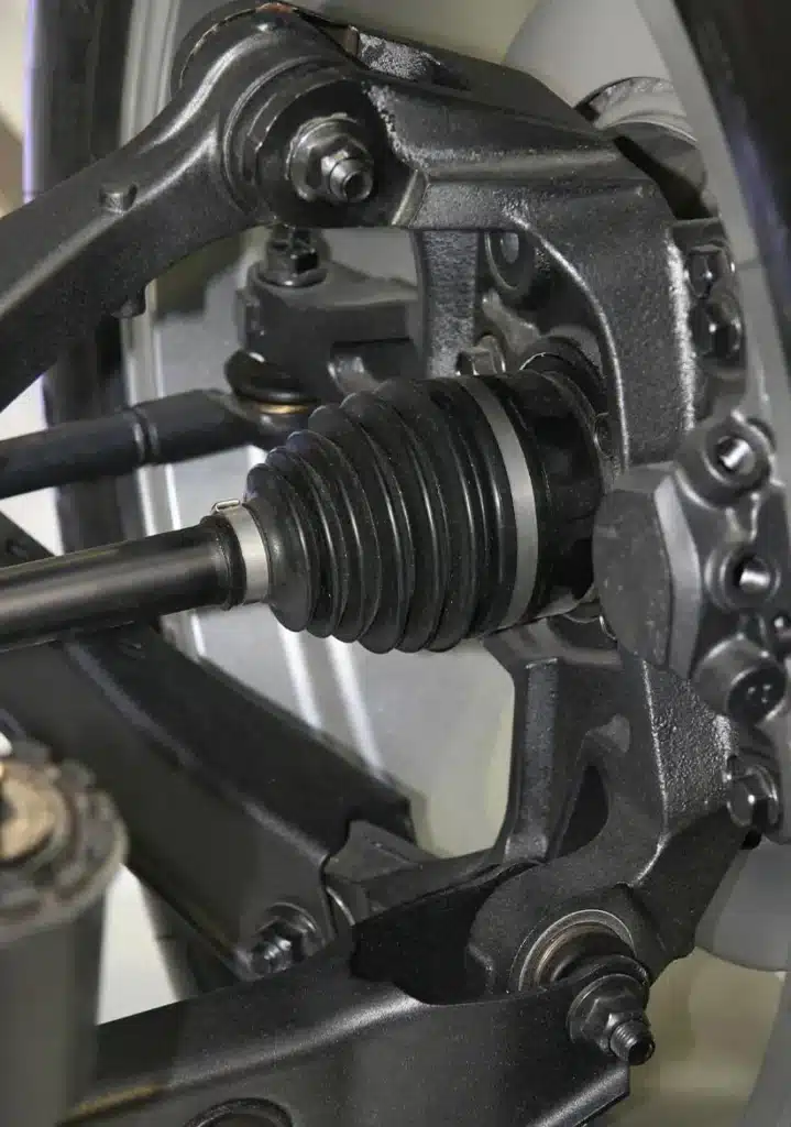 Axle Repair & Service in Clive, IA At Westside Auto Pros. Close-up of a vehicle’s suspension system showing a constant velocity (CV) joint, black rubber boot, metal components, bolts, and part of a wheel rim in the background.