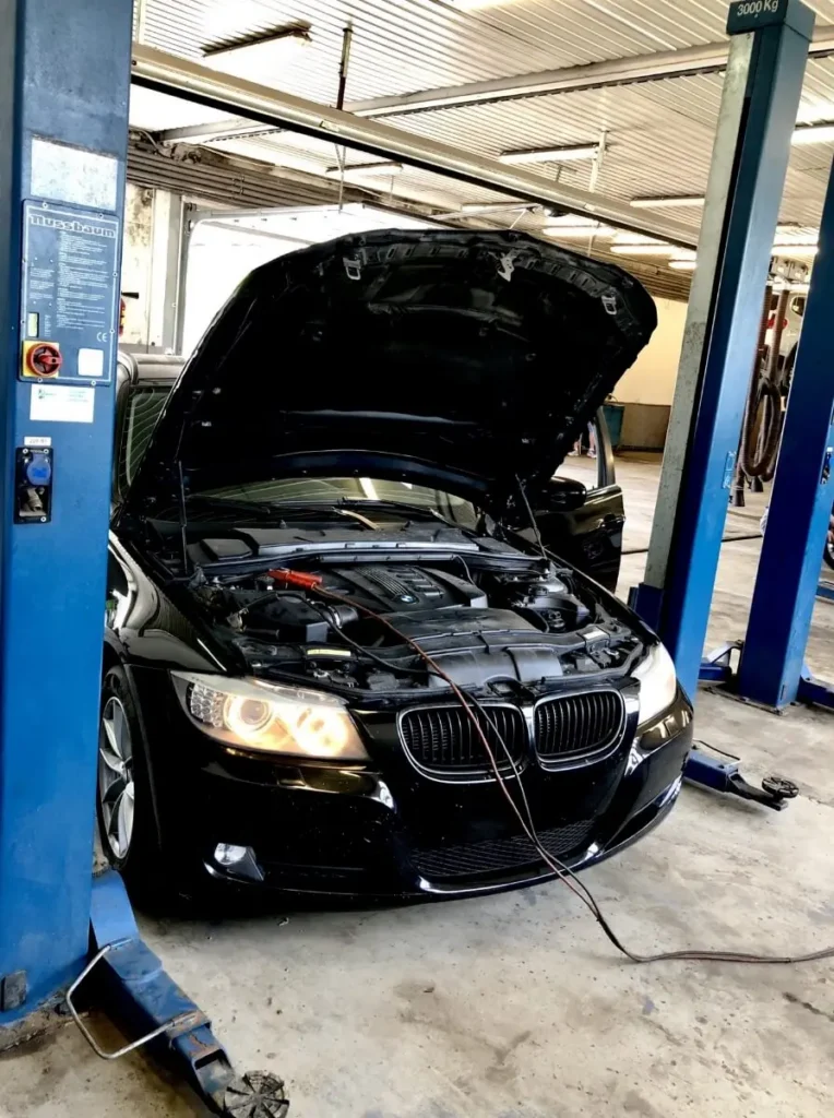 Vehicle Diagnostics in Clive, IA At Westside Auto Pros. A black car with its hood open is parked in a garage between blue lifts, with jumper cables attached to the engine. The car’s headlights are on and the workshop is well-lit.