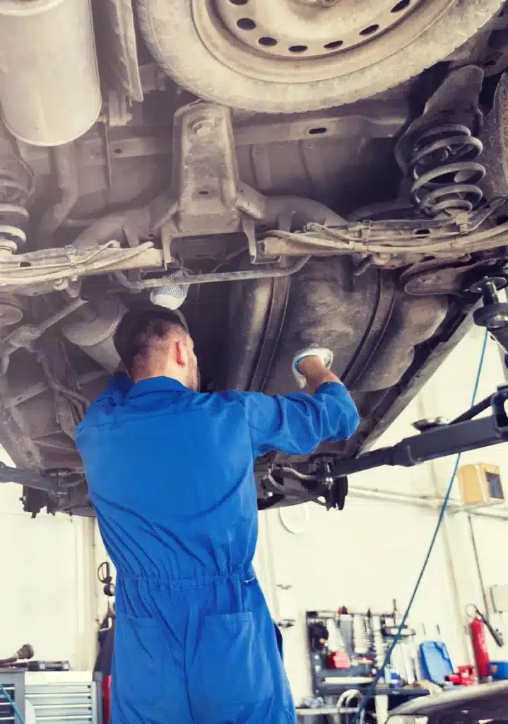 Drivetrain Expert in Clive, IA At Westside Auto Pros. A mechanic in a blue jumpsuit works underneath a lifted car in an auto repair shop, inspecting or repairing the vehicle’s undercarriage. Various tools and equipment are visible in the background.