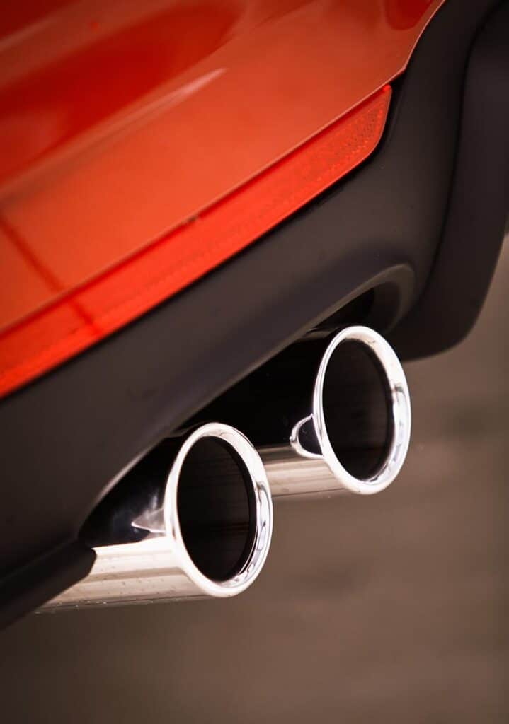 Exhaust Repairs in Clive, IA At Westside Auto Pros. Close-up view of a car’s dual chrome exhaust pipes beneath a red bumper, with part of a black trim and a red reflector visible.