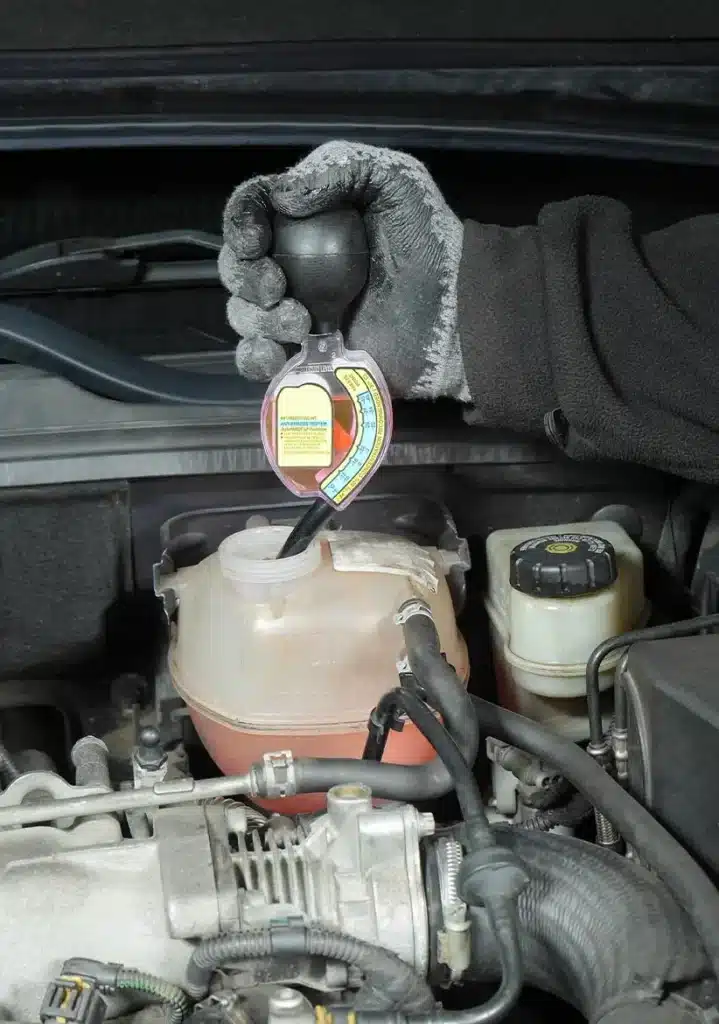 Brake Fluid, Fluid Services in Clive, IA At Westside Auto Pros. A gloved hand uses a coolant tester to check the antifreeze level in a cars coolant reservoir under the hood. Engine components and hoses are visible around the reservoir.