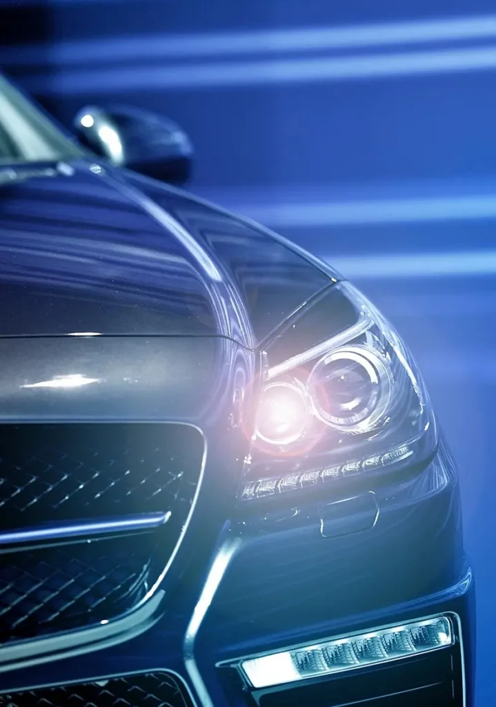 Headlight Replacement in Clive, IA At Westside Auto Pros. Close-up of the front of a dark-colored luxury car with its headlights on, set against a blue background with horizontal light streaks.