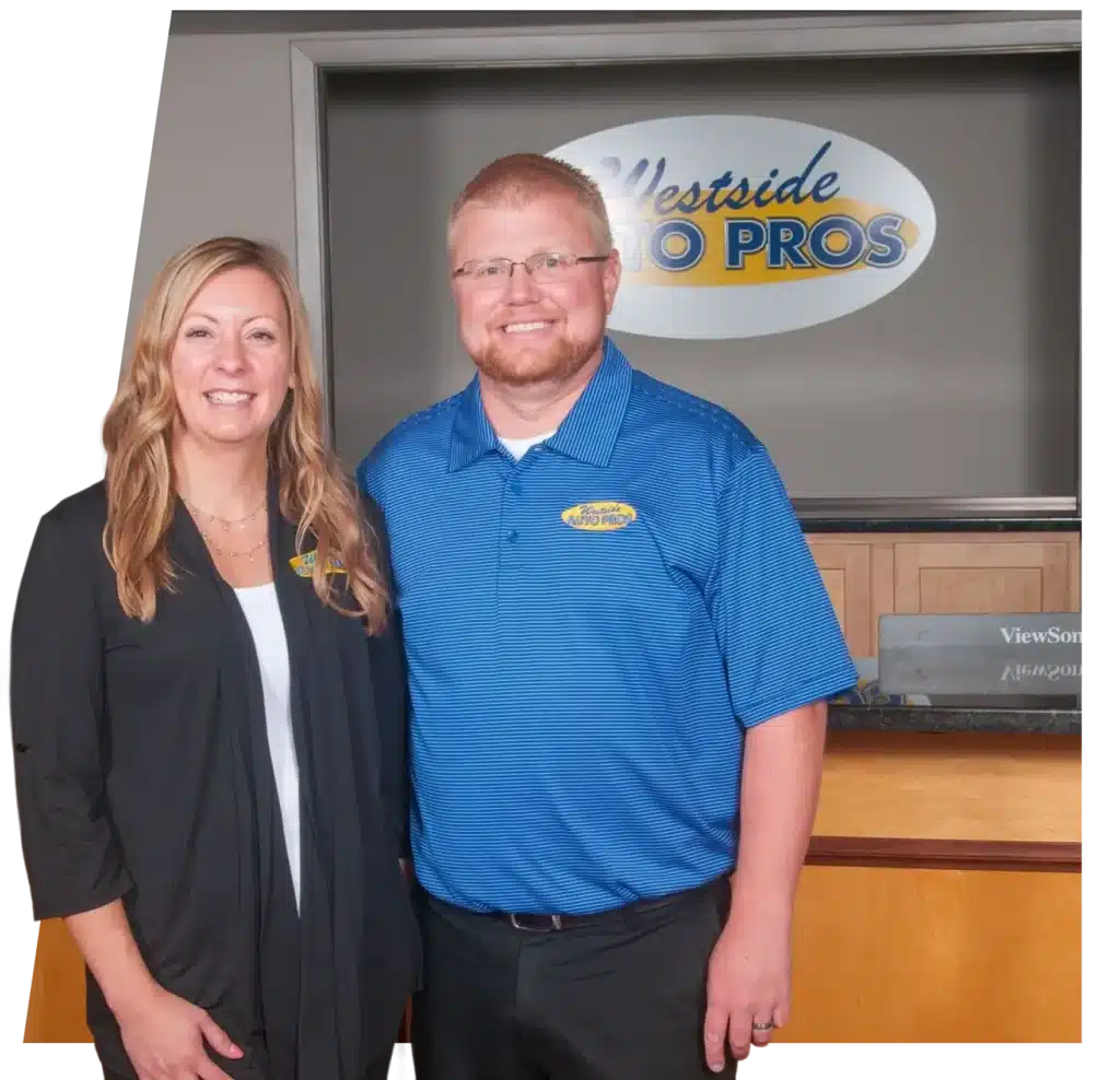 About Westside Auto Pros in Clive, IA. A woman and a man stand smiling in front of a reception desk with a Westside Auto Pros sign on the wall behind them. Both are wearing business attire with company logos.