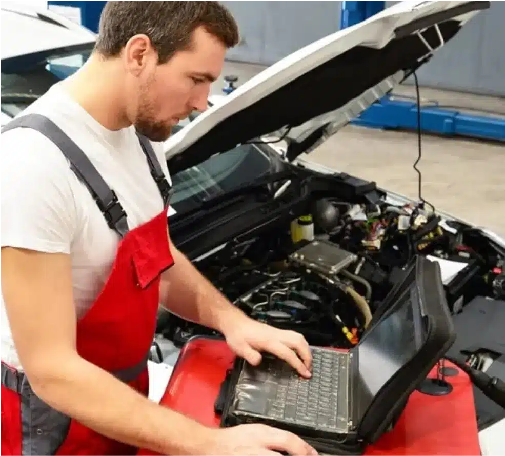 Auto Diagnostics in Clive, IA At Westside Auto Pros. A mechanic in red overalls uses a laptop computer while inspecting the engine of a car with its hood open in a garage or workshop.