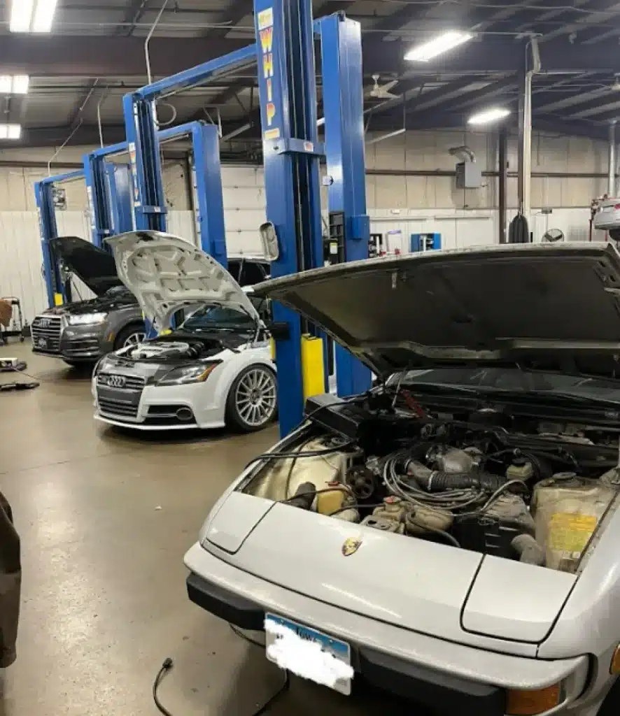 Auto Repair Services in Clive, IA At Westside Auto Pros. Three cars with open hoods are parked in a repair shop with blue hydraulic lifts; the closest car is a white Porsche, and the other two appear to be Audis. The workshop is spacious with a high ceiling.