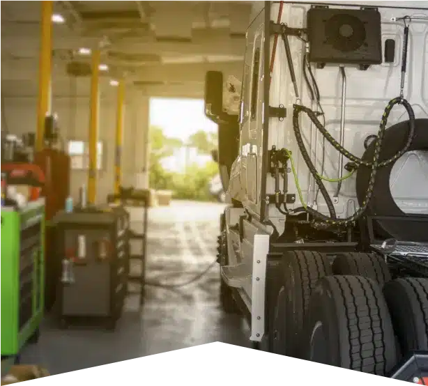 Diesel Repair in Clive, IA At Westside Auto Pros. A white semi-truck parked inside a brightly lit garage or workshop, with visible tools and equipment on shelves and carts nearby. The image focuses on the rear side of the truck.