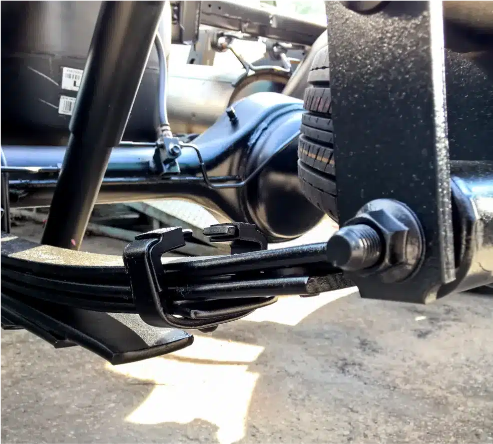 Suspension & Steering Repair in Clive, IA At . Westside Auto ProsClose-up view of a vehicles black leaf spring suspension system, showing metal leaf springs, bolts, brackets, and part of a tire in the background, all set against a concrete ground.