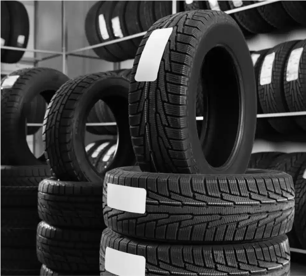Tire Sales & Services in Clive, IA At Westside Auto Pros. Stacks of car tires with white labels are arranged in rows inside a tire shop. Shelves filled with more tires are visible in the background. The image is in black and white.