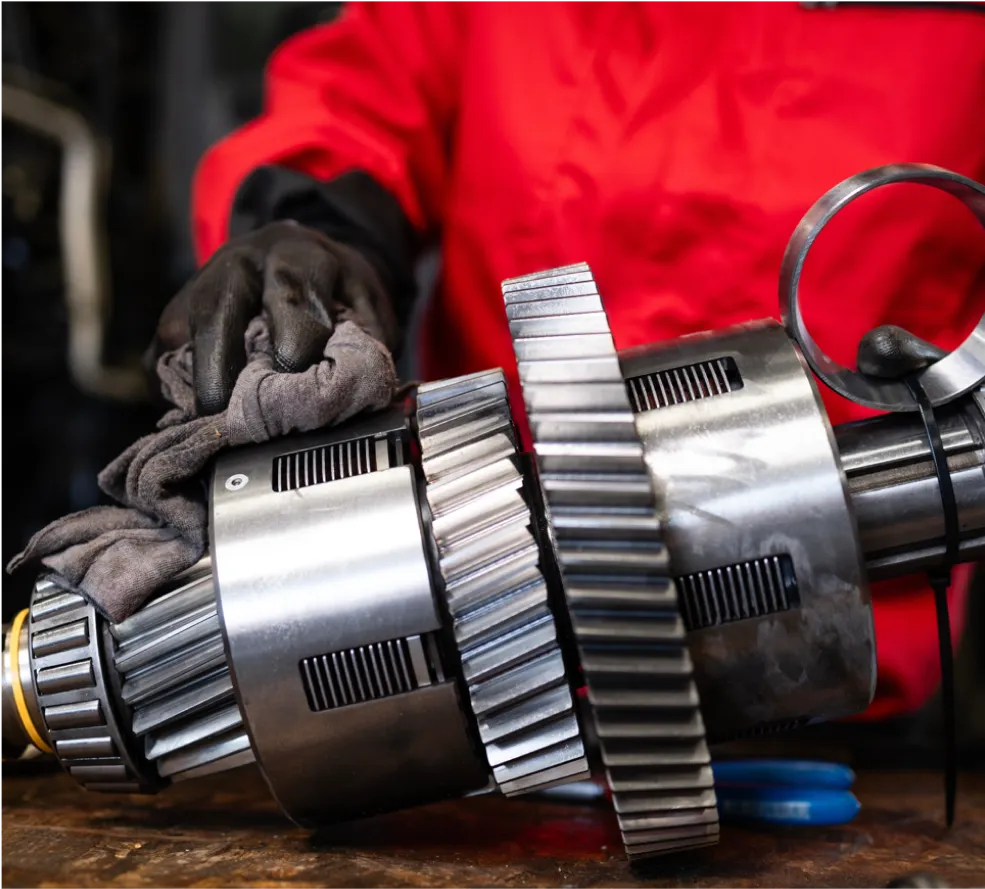 Transmission Repair & Replacement in Clive, IA At Westside Auto Pros. A person wearing black gloves and a red jacket is cleaning a large metal gear mechanism with a cloth on a workbench.