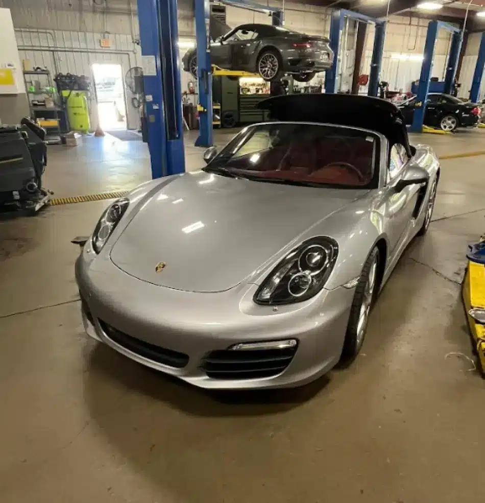 Vehicles We Services in Clive, IA At Westside Auto Pros. A silver Porsche convertible with its top partially open is parked inside an auto repair shop. In the background, another black Porsche is elevated on a hydraulic lift.