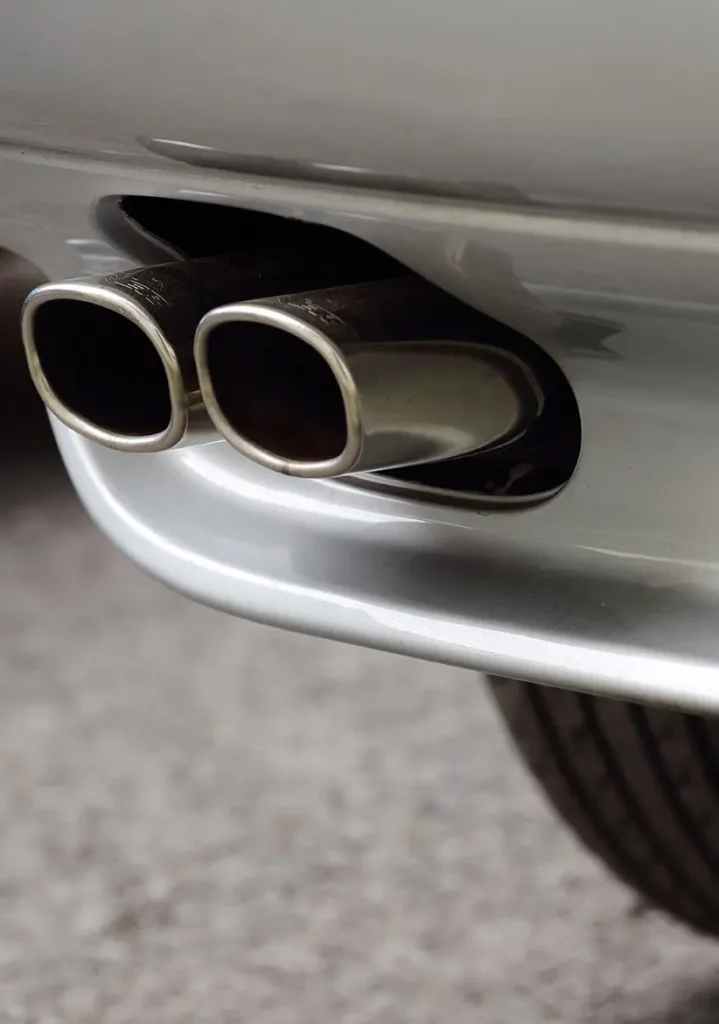 Muffler Repair Services in Clive, IA At Westside Auto Pros. Close-up of a car’s dual exhaust pipes with a shiny metallic finish, extending from the rear bumper. The car’s silver body and a portion of the rear tire are visible in the background.