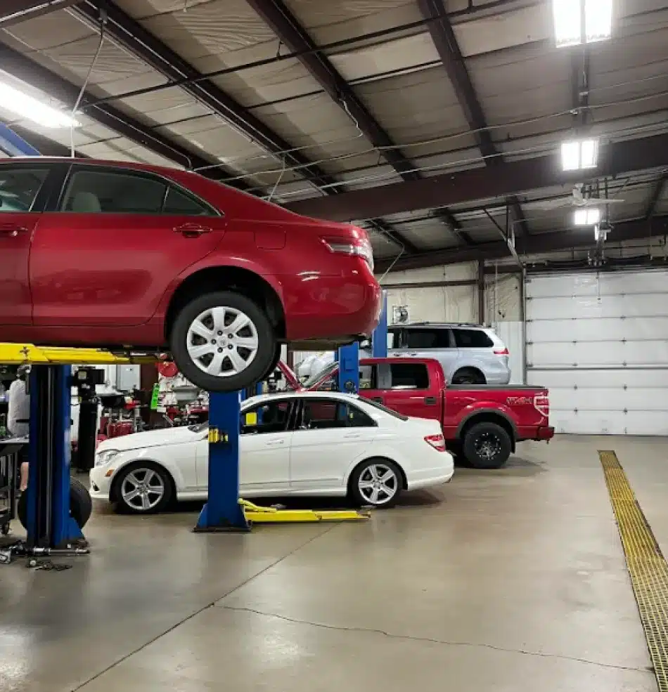 A red car is lifted on a hydraulic platform in an auto repair shop specializing in asian vehicle repair, with a white car and a red truck parked nearby. Mechanics and tools are visible in the spacious, well-lit garage.