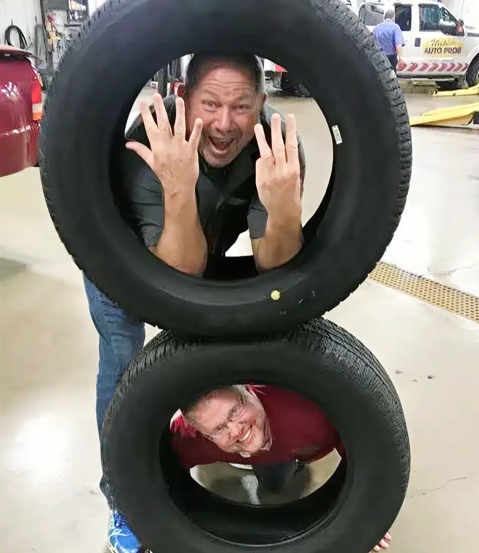 Two men pose playfully inside stacked tires in a Clive IA auto shop; one at the top smiles and holds up seven fingers, while the other crouches in the bottom tire and grins at the camera. Perfect snapshot of fun at work in auto repair careers.