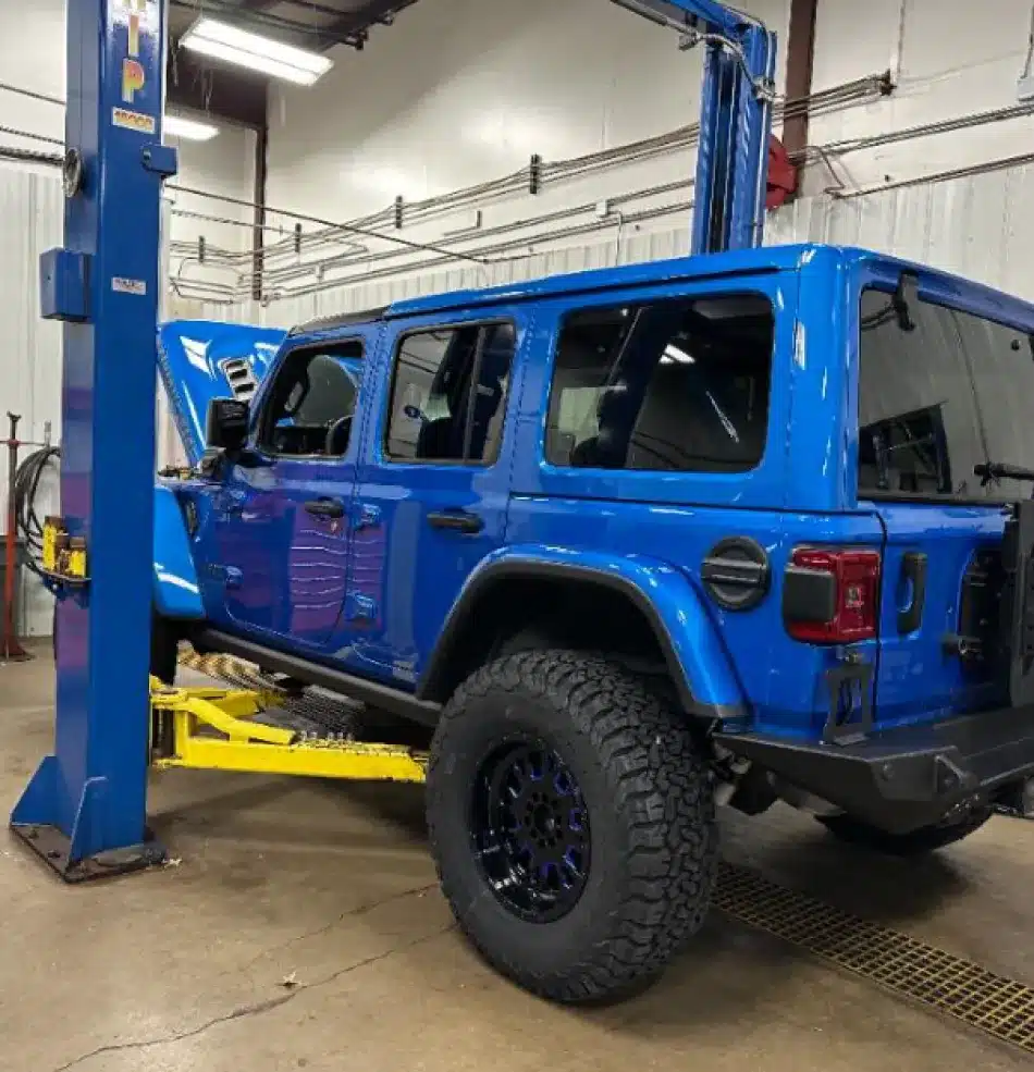 A blue Jeep Wrangler is lifted on a hydraulic car lift inside a garage with its hood open, showing it is undergoing domestic vehicle repair by ASE certified service technicians.