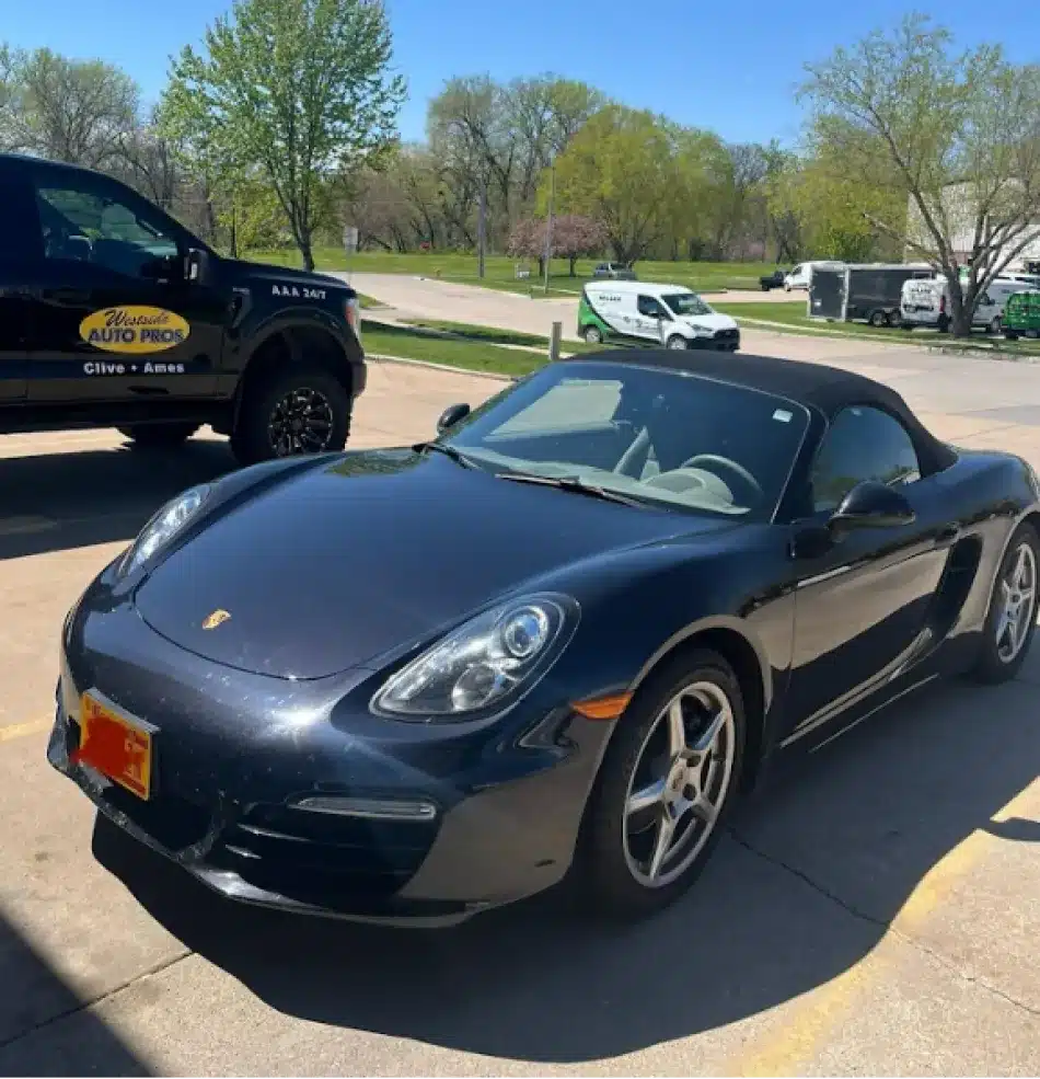 A black Porsche convertible, symbolizing luxury car service, is parked outside on a sunny day near a black pickup truck with “Auto Pros” signage. Trees and other vehicles are visible in the background.