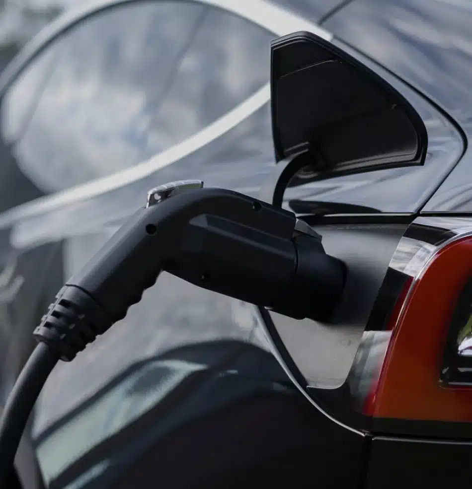 A close-up of an electric car being charged, showing the charging cable plugged in next to the rear taillight. Perfect for showcasing hybrid & EV vehicle repair expertise. The side mirror is folded, reflecting the sky on the car’s surface.